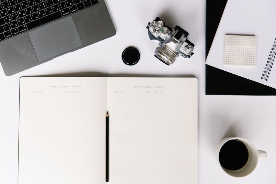 Flat lay of workspace with open notebook, coffee, camera, laptop, and stationery for creative work.