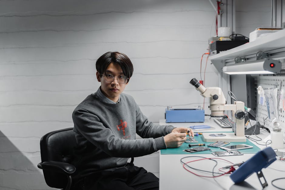 Asian male technician working with electronics in a lab setting, focused on a microscope and devices.
